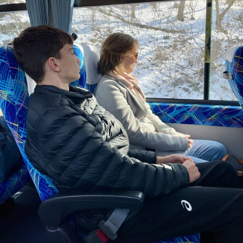 Two people sitting on the Nagano bus to Hakuba, looking out the window and seeing snow on side of road.