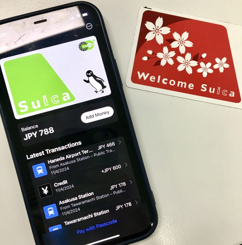 An image of a mobile phone with the Japan Suica card transportation app on screen, sitting next to a physical transportation card of the 'Welcome Suica' card